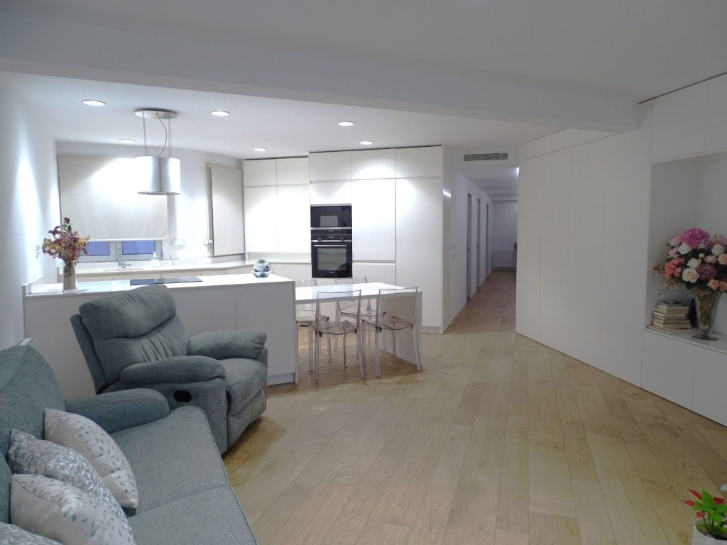 Sala de estar abierta con cocina, sofá, mesa con sillas transparentes y flores decorativas.