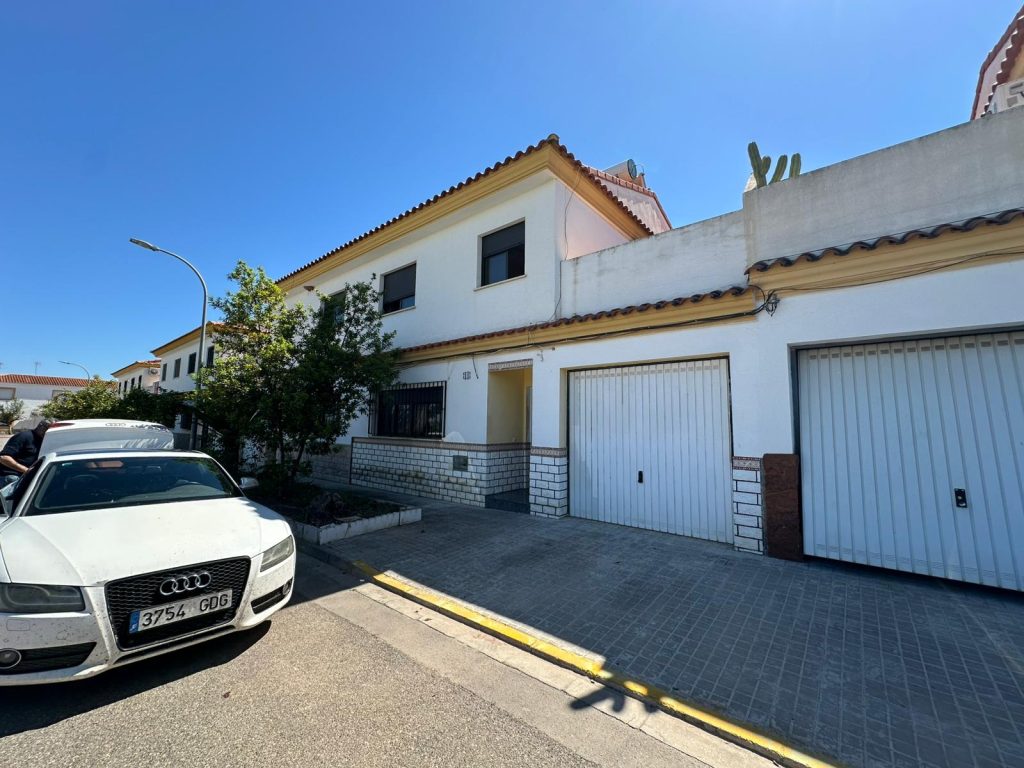 Casa blanca de dos pisos con dos garajes y jardín pequeño, coche blanco en la calle frente.